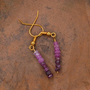 May include: A pair of dangle earrings featuring gold-tone ear wires and small gold beads. Each earring consists of a vertical strand of faceted gemstone beads that transition in colour from light pink at the top to deep purple at the bottom.
