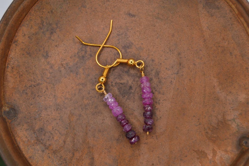 May include: A pair of dangle earrings featuring gold-tone ear wires and small gold beads. Each earring consists of a vertical strand of faceted gemstone beads that transition in colour from light pink at the top to deep purple at the bottom.