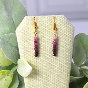 May include: A pair of drop earrings featuring gold-toned hooks and a vertical arrangement of small, faceted beads in a gradient of pink and deep magenta hues. The beads are strung in a linear pattern, transitioning from lighter pink to dark purple.