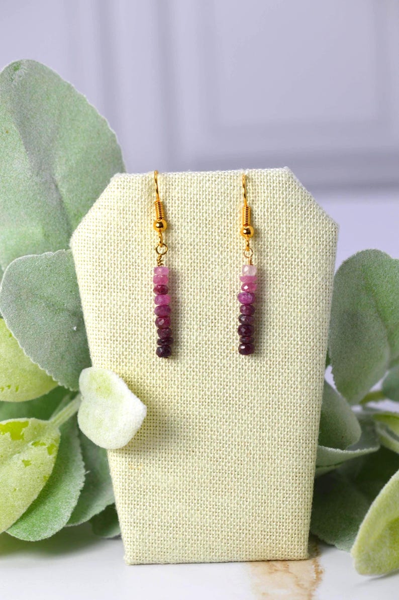 May include: A pair of drop earrings featuring gold-toned hooks and a vertical arrangement of small, faceted beads in a gradient of pink and deep magenta hues. The beads are strung in a linear pattern, transitioning from lighter pink to dark purple.