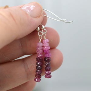 May include: Dangle earrings featuring a vertical strand of faceted, round, pink-to-deep-red ombre gemstone beads suspended from silver-toned metal ear wires. The beads transition in colour from light pink at the top to dark burgundy at the bottom.