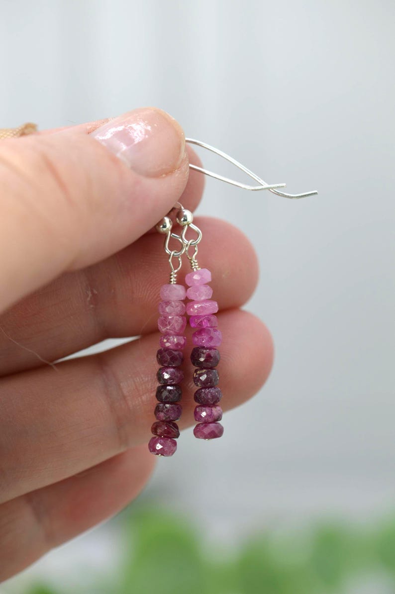 May include: Dangle earrings featuring a vertical strand of faceted, round, pink-to-deep-red ombre gemstone beads suspended from silver-toned metal ear wires. The beads transition in colour from light pink at the top to dark burgundy at the bottom.