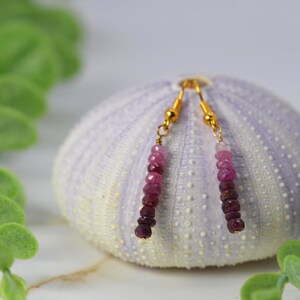 May include: A pair of drop earrings featuring faceted, round beads in a gradient of pink to deep burgundy red. The beads are strung on thin gold-toned metal wire with gold-toned ear hooks, creating a delicate and slender vertical drop design.