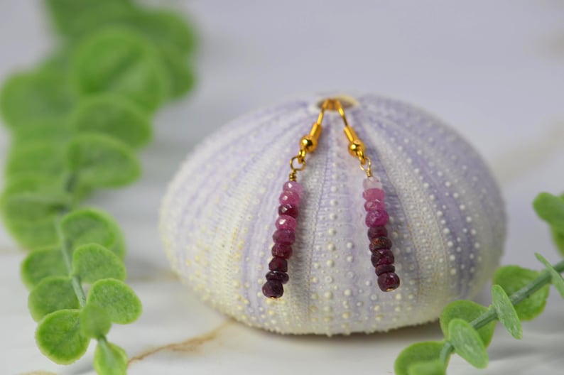 May include: A pair of drop earrings featuring faceted, round beads in a gradient of pink to deep burgundy red. The beads are strung on thin gold-toned metal wire with gold-toned ear hooks, creating a delicate and slender vertical drop design.