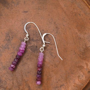 May include: A pair of drop earrings featuring a vertical arrangement of small, faceted gemstone beads in a gradient of pink, magenta, and deep purple hues. The beads are suspended from silver-toned metal ear wires with a simple hook design.