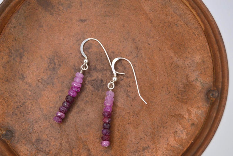 May include: A pair of drop earrings featuring a vertical arrangement of small, faceted gemstone beads in a gradient of pink, magenta, and deep purple hues. The beads are suspended from silver-toned metal ear wires with a simple hook design.