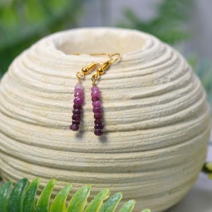 May include: Dangle earrings featuring a vertical row of small, faceted gemstone beads in an ombre colour gradient, transitioning from light pink at the top to deep purple at the bottom, suspended from gold-toned metal ear wires.