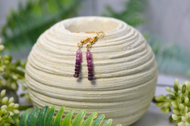 May include: Dangle earrings featuring a vertical row of small, faceted gemstone beads in an ombre colour gradient, transitioning from light pink at the top to deep purple at the bottom, suspended from gold-toned metal ear wires.