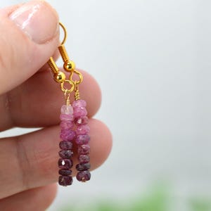 May include: A pair of drop earrings featuring faceted gemstone beads in a vertical ombre gradient, transitioning from light pink at the top to deep dark purple at the bottom, suspended from gold-toned metal ear wires with a simple hook design.