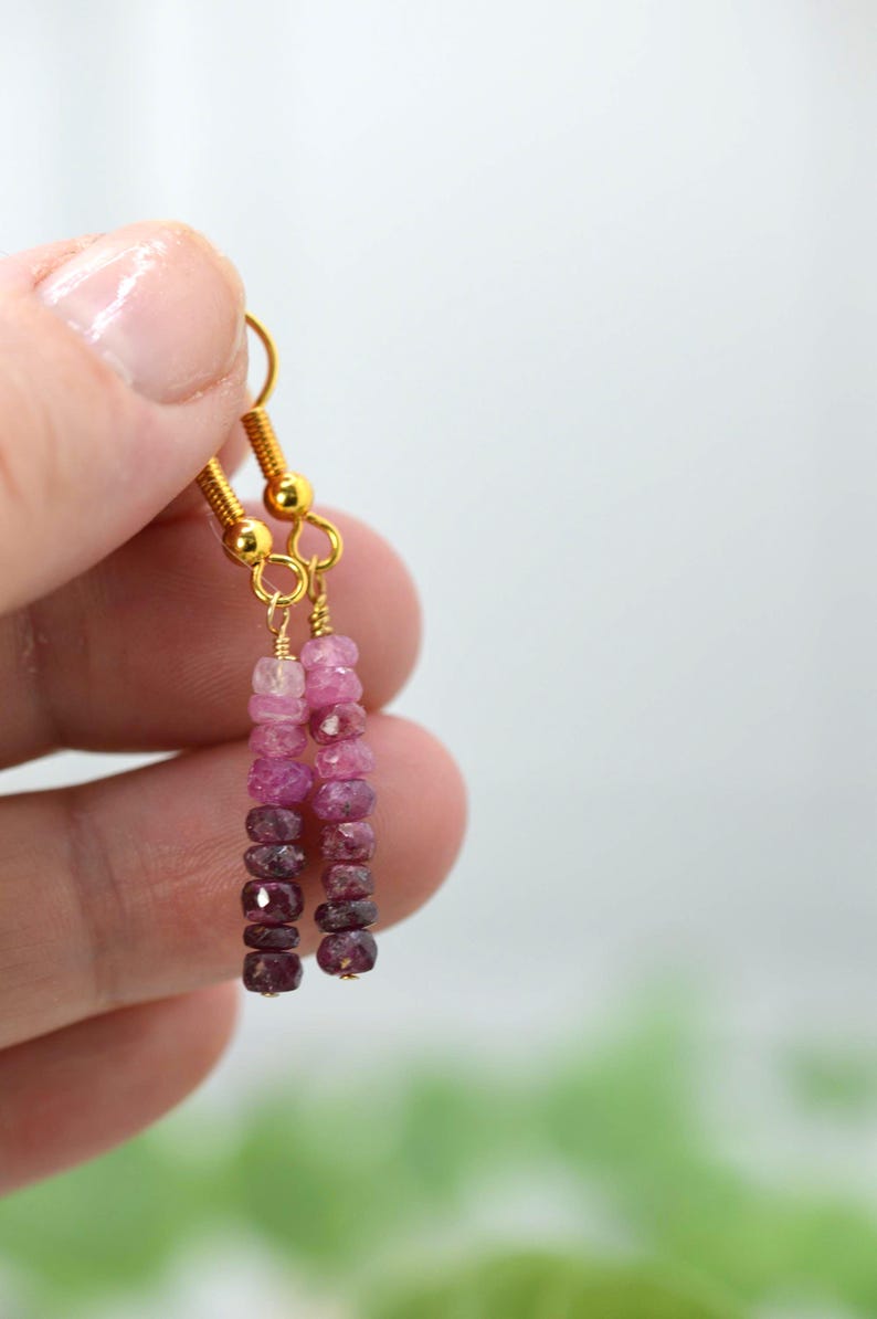 May include: A pair of drop earrings featuring faceted gemstone beads in a vertical ombre gradient, transitioning from light pink at the top to deep dark purple at the bottom, suspended from gold-toned metal ear wires with a simple hook design.