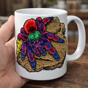 May include: White ceramic mug featuring a colorful illustration of a tarantula spider. The spider has a red, green, purple, and blue design, set against a brown, textured background. The mug has a curved handle.