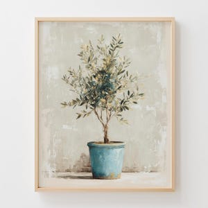 Op de afbeelding: Een ingelijste print van een olijfboom in een blauwe pot. De boom heeft groene en witte bladeren en een bruine stam. De achtergrond is een getextureerd, gedempt groen en beige. Het frame is licht van kleur.