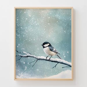 May include: A framed art print depicting a small bird with black and white plumage, perched on a snow-laden branch. The background is a soft blue and white winter scene with falling snow. The artwork is presented in a wooden frame.