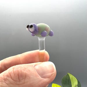 May include: A glass turtle figurine with purple and green accents and large black eyes. The turtle is perched on a clear glass rod.