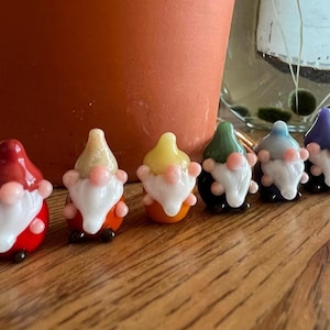 Może przedstawiać: Zestaw ośmiu małych szklanych krasnoludków, każdy z innym kolorowym kapeluszem i białą brodą. Krasnoludki są ułożone w rzędzie na drewnianej powierzchni.