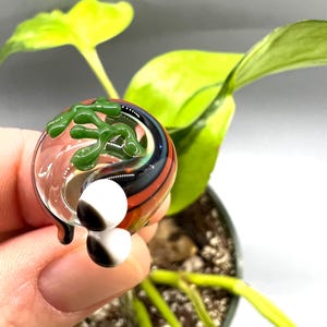 Puede incluir: Un globo de vidrio transparente con detalles verdes, negros y naranjas, con dos esferas blancas y negras. El globo tiene un diseño orgánico verde en la parte superior. El fondo incluye hojas verdes y una pequeña planta en maceta.