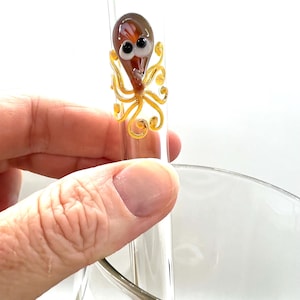 Op de afbeelding: Een heldere glazen steel met een klein, kleurrijk glazen octopusbeeldje bevestigd. De octopus heeft een bruine kop, witte ogen en gele tentakels. Het beeldje is bevestigd aan een heldere glazen steel.