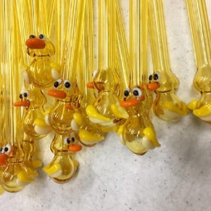 May include: A group of yellow glass drinking straws shaped like ducks with orange eyes and beaks. The straws are hanging from a string.