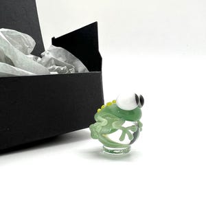 May include: A small green glass figurine of a frog with large white eyes and a yellow stripe on its back. The frog is sitting on a clear glass base.