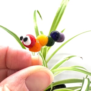 May include: A colorful glass caterpillar figurine with a red head, orange body, yellow, green, blue, and purple segments. The caterpillar has black and white eyes and is held in front of a plant with green and white striped leaves.
