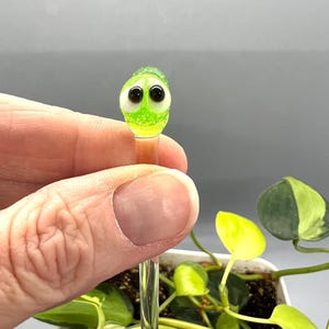 May include: A green glass figurine with googly eyes on a clear glass rod. The figurine is being held by a hand and is in front of a potted plant.