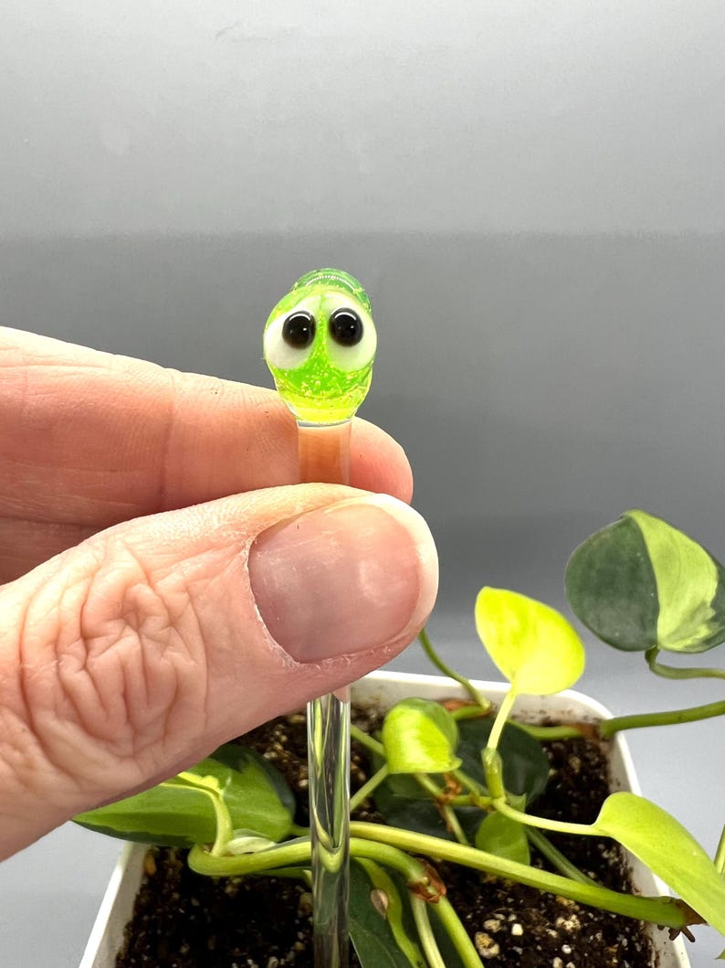 May include: A green glass figurine with googly eyes on a clear glass rod. The figurine is being held by a hand and is in front of a potted plant.