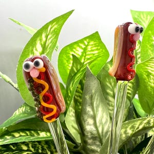 May include: Glass hot dog food picks with googly eyes, red ketchup, yellow mustard, and green relish. The hot dogs are on clear glass stems, set against a backdrop of green leaves.