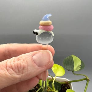 May include: A glass plant stake with a whimsical, colorful, and layered glass topper. The topper features a blue hat, pink, beige, and white layers, and two black eyes.