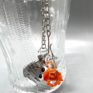 Op de afbeelding: Een zilveren thee-ei met een ketting en een kleine oranje glazen krab-bedel. Het thee-ei hangt aan de rand van een heldere glazen mok.