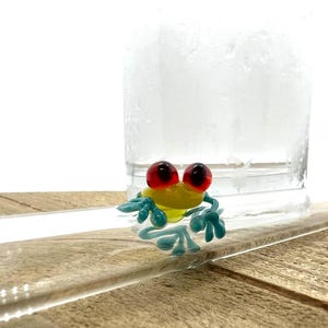 May include: A small glass frog figurine featuring a bright yellow body, translucent teal limbs with detailed toes, and two prominent, spherical red eyes. The frog is positioned on a clear, curved glass surface, showcasing its smooth texture.