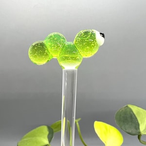 May include: A green glass worm with a white eye, attached to a clear glass rod. The worm is made of multiple round, connected pieces.