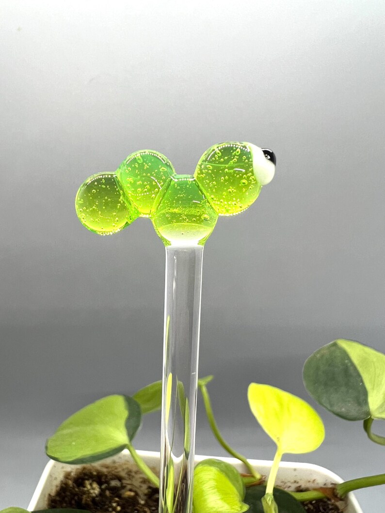May include: A green glass worm with a white eye, attached to a clear glass rod. The worm is made of multiple round, connected pieces.