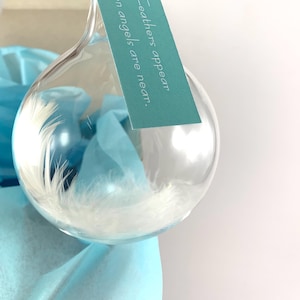 May include: A clear glass ornament filled with white feathers and light blue tissue paper. A teal tag is attached with the text "Feathers appear when angels are near."