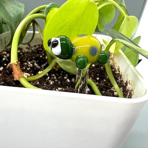 May include: A glass turtle plant stake with a yellow body and green head and legs. The turtle has blue spots and black and white eyes. The stake is placed in a white planter with green plants.