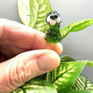 May include: A green glass figurine of a cartoon-like creature with black spots, white eyes, and a pink bubble. The figurine is held in front of green and yellow leaves.