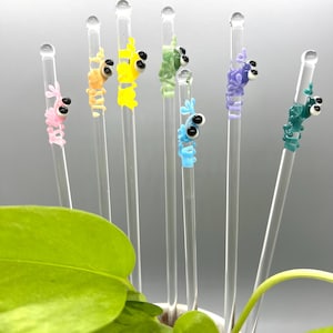 May include: A collection of colorful glass drink stirrers. Each stirrer features a whimsical, cartoon-like creature in shades of pink, orange, yellow, green, blue, purple, and teal. The stirrers are clear with a rounded top.