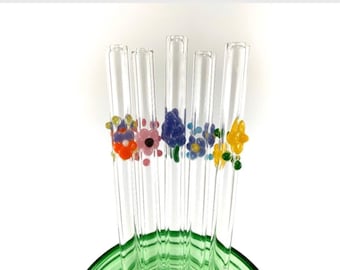 May include: Set of six glass drinking straws with colorful flower designs. The straws are clear glass with a variety of colored flowers, including purple, orange, yellow, and green.