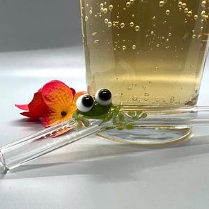 以下が含まれることがあります： 透明なガラス製のストローに、緑色のカエルの置物が付いている。カエルは黒い瞳の白い目をしており、緑色の体をしている。