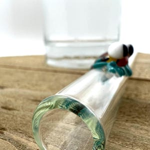 May include: A clear glass drinking straw featuring a small, decorative glass figurine of a creature with large white eyes, a red nose, and teal limbs attached to the side. The straw is cylindrical with a smooth, transparent finish throughout.