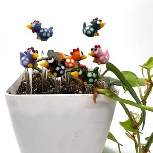 May include: A set of eight colorful glass chicken figurines with white polka dots. The figurines are arranged in a white planter with soil and a green plant.