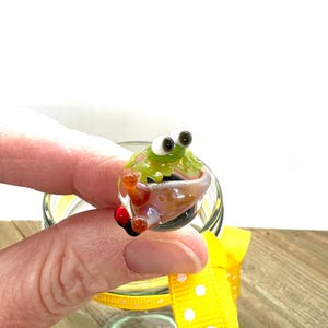 May include: A miniature glass frog with large black eyes and a green body sits in a tiny pot. The frog is held above a clear glass jar with a yellow ribbon tied around it. The ribbon has white polka dots.