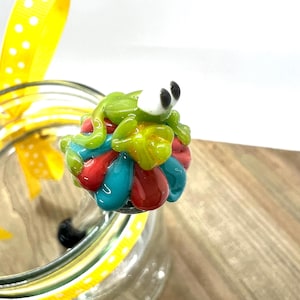 May include: A glass jar with a decorative frog lid. The frog is green with white eyes and sits atop a cluster of red, blue, and yellow glass beads. A yellow ribbon with white polka dots is tied to the jar.