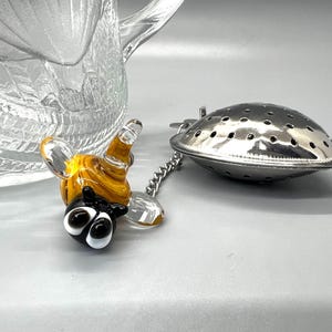 May include: A glass bee tea infuser with a stainless steel mesh tea strainer. The bee is yellow and black with large eyes. The strainer is attached to a chain.