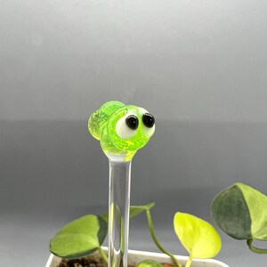 May include: A green glass bug with black eyes on a clear glass stick. The bug is sitting on top of a potted plant.