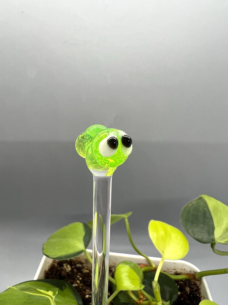 May include: A green glass bug with black eyes on a clear glass stick. The bug is sitting on top of a potted plant.