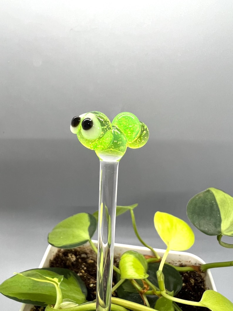 May include: A green glass bug with black eyes and white pupils on a clear glass rod. The bug is sitting on top of a potted plant.