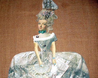Puede incluir: Muñeca de papel de una mujer con un vestido de época. La muñeca tiene un vestido azul claro con un patrón de encaje blanco, un gorro a juego y un abanico. La muñeca sostiene un pequeño libro. El fondo es marrón texturizado.