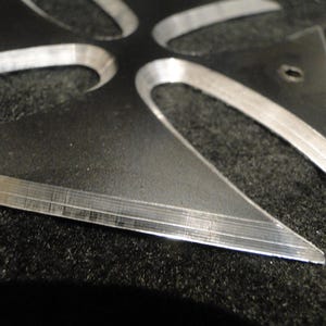 Puede incluir: Una cruz de metal plateado con un dise&ntilde;o afilado y puntiagudo. La cruz est&aacute; hecha de dos capas de metal, creando un efecto de capas.