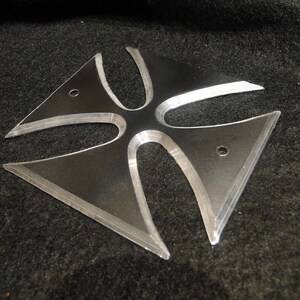 Puede incluir: Una cruz de metal plateado con cuatro puntas, cada punta tiene un borde redondeado. La cruz tiene un centro elevado y una superficie ligeramente texturizada.