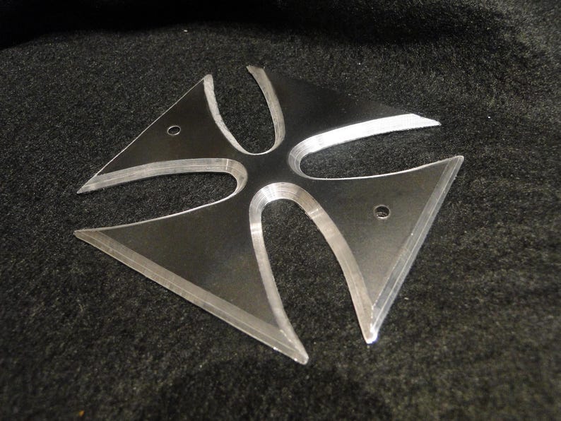 Puede incluir: Una cruz de metal plateado con cuatro puntas, cada punta tiene un borde redondeado. La cruz tiene un centro elevado y una superficie ligeramente texturizada.